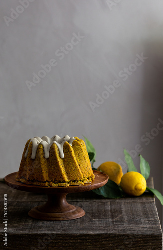 Yellow lemon turmeric cake with white chocolate glazing served on a wooden cake stand on a wooden table with fresh lemons and lemon leaves on the side. 