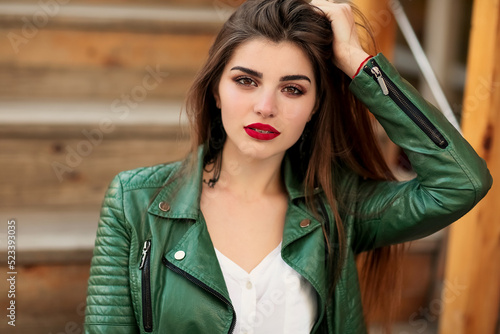 Fashion outdoor portrait of gorgeous long hair woman in green leather jacket and red lips, autumn style. Fashionable hipster girl in trendy casual clothes posing at city street lifestyle.