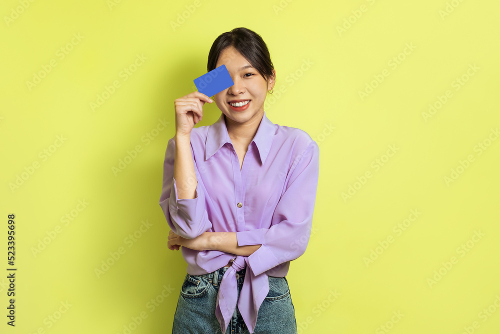 Cheerful Korean Female Holding Credit Card Near Face, Yellow Background Cheerful Korean Female Holding Credit Card Near Face, Yellow Background