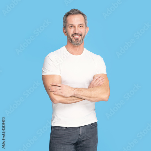 Handsome middle aged grey haired man standing with arms folded happy looking at camera wearing white t-shirt and jeans isolated on blue background. Mature fit man, healthy lifestyle concept