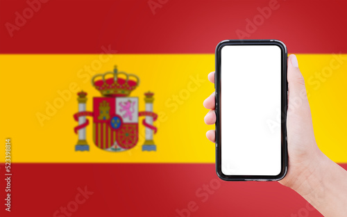 Close-up of male hand holding smartphone with blank on screen, on background of blurred flag of Spain.
