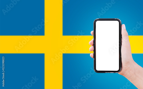 Close-up of male hand holding smartphone with blank on screen, on background of blurred flag of Sweden.