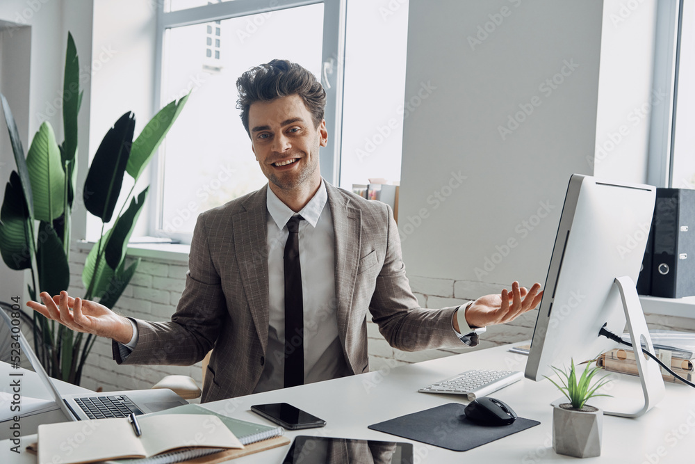 Naklejka premium Cheerful young man in formalwear looking at camera and gesturing while sitting in office
