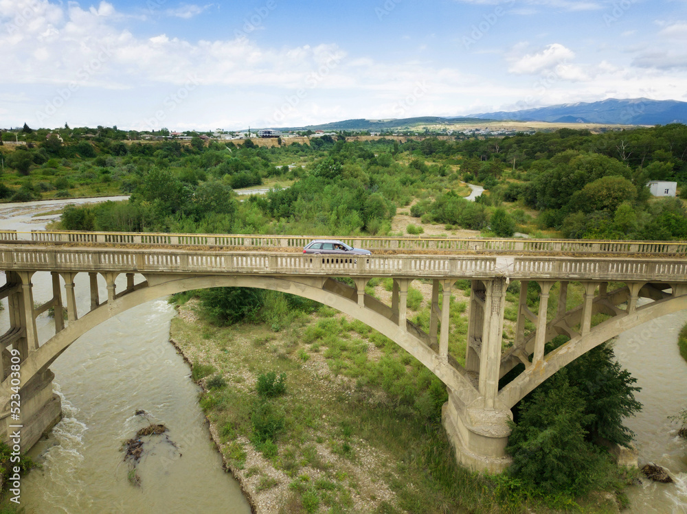Fototapeta premium Car rides on an old arch bridge over a mountain river on a summer day, a side view from a drone