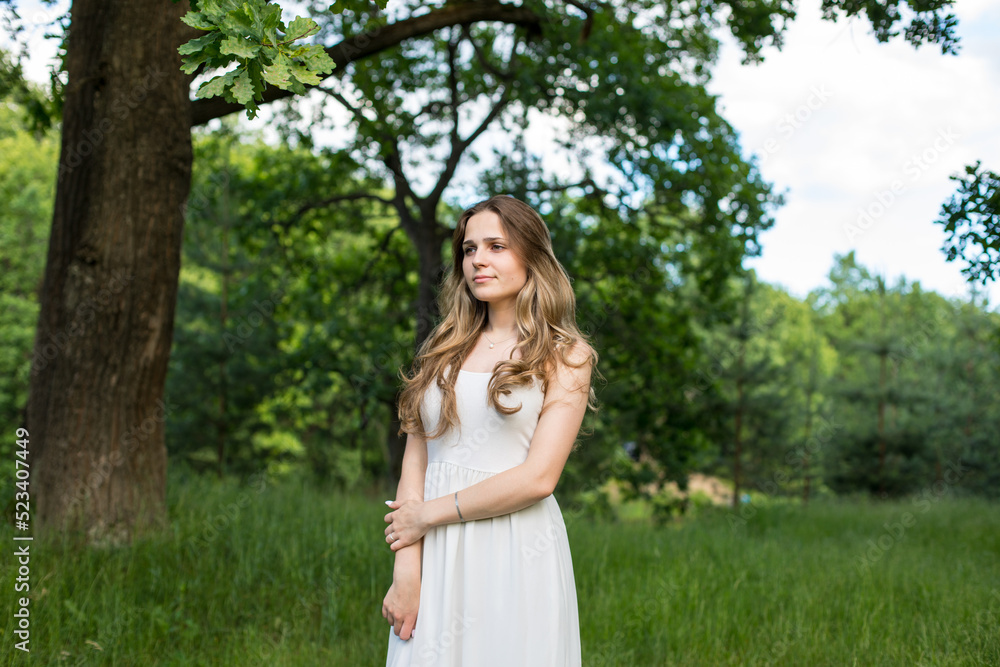 Beautiful young Russian girl dressed in a white dress, walks through the forest among the oaks in the rays of the sunset Beautiful young Russian girl dressed in a white dress, walks through the forest among the oaks in the rays of the sunset