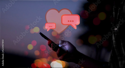 illustrated red speech bubbles in front of a heart above a hand holding a smartphone with screen glowing at night, red, orange and yellow dots of light, romantic, dating, flirt, love photo
