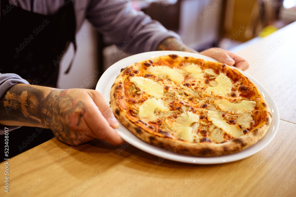 Faceless view of tattooed cook holding delicious hawaiian pizza in pizzeria 