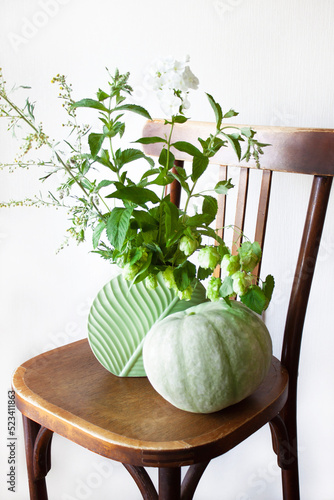 Autumn still life. A green bouquet in a vase of wormwood, mint, hops and phlox sprigs. A green pumpkin and a vase of flowers on a chair. Harvesting. Healthy lifestyle. Boho style