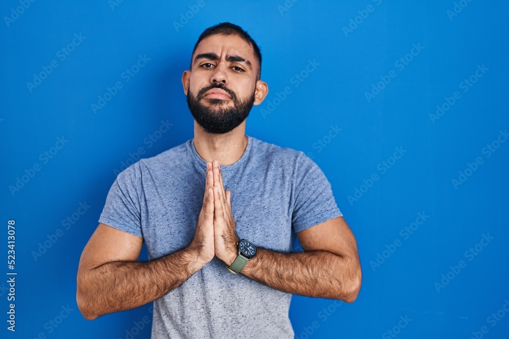 Middle east man with beard standing over blue background begging and praying with hands together with hope expression on face very emotional and worried. begging. Middle east man with beard standing over blue background begging and praying with hands together with hope expression on face very emotional and worried. begging.