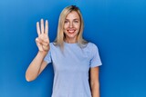 Beautiful blonde woman wearing casual t shirt over blue background showing and pointing up with fingers number three while smiling confident and happy.