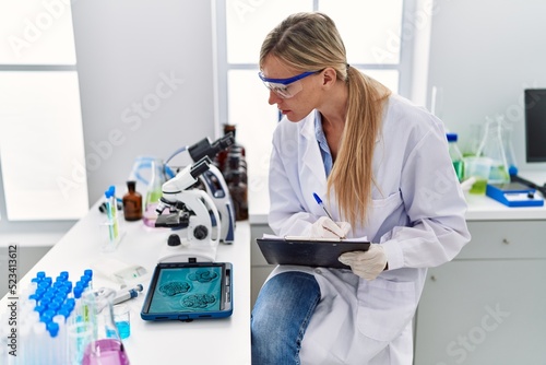 Young blonde woman wearing scientist uniform writing on clipboard looking embryion images at laboratory photo