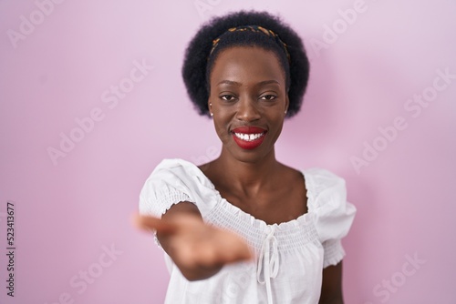African woman with curly hair standing over pink background smiling cheerful offering palm hand giving assistance and acceptance.