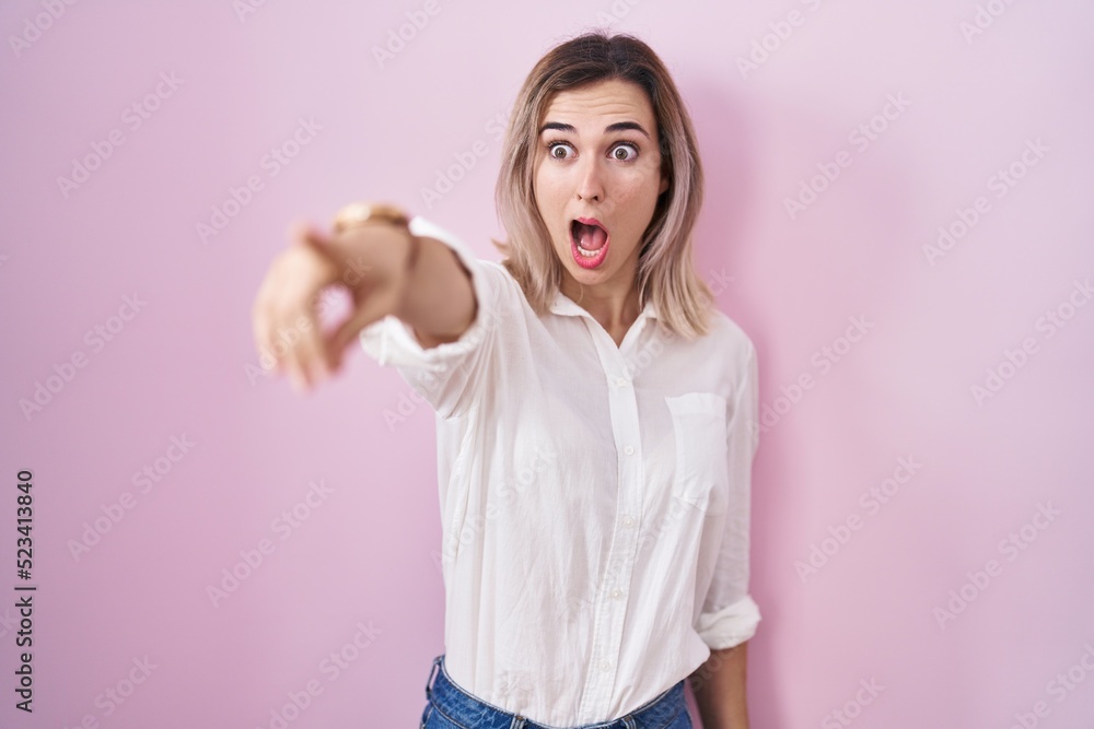 Young beautiful woman standing over pink background pointing with finger surprised ahead, open mouth amazed expression, something on the front