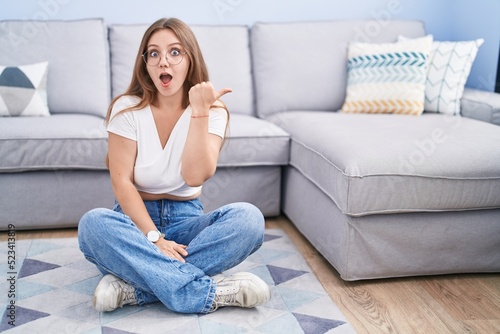 Young caucasian woman sitting on the floor at the living room surprised pointing with hand finger to the side, open mouth amazed expression.