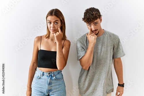 Young beautiful couple standing together over isolated background pointing to the eye watching you gesture, suspicious expression