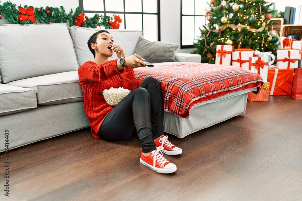 Obraz premium Young hispanic woman watching movie sitting by christmas tree at home