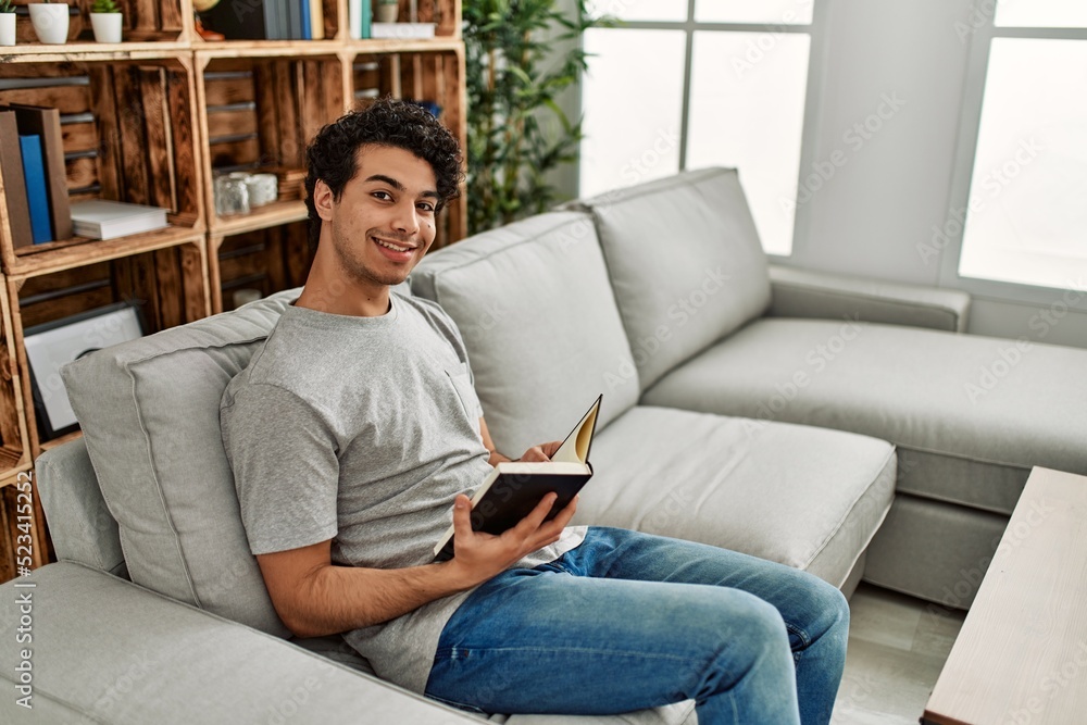 Young hispanic man reading book sitting on the sofa at home. Young hispanic man reading book sitting on the sofa at home.