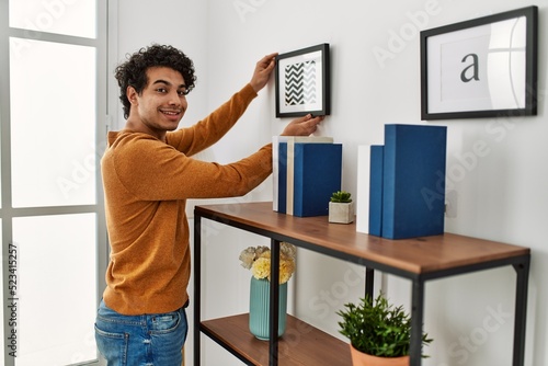 Young hispanic man smiling happy hanging frame on wall at home.