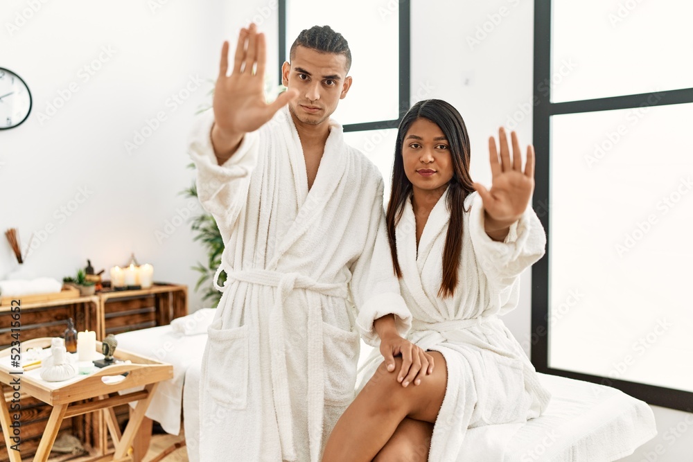 Young latin couple wearing towel standing at beauty center doing stop sing with palm of the hand. warning expression with negative and serious gesture on the face. Young latin couple wearing towel standing at beauty center doing stop sing with palm of the hand. warning expression with negative and serious gesture on the face.