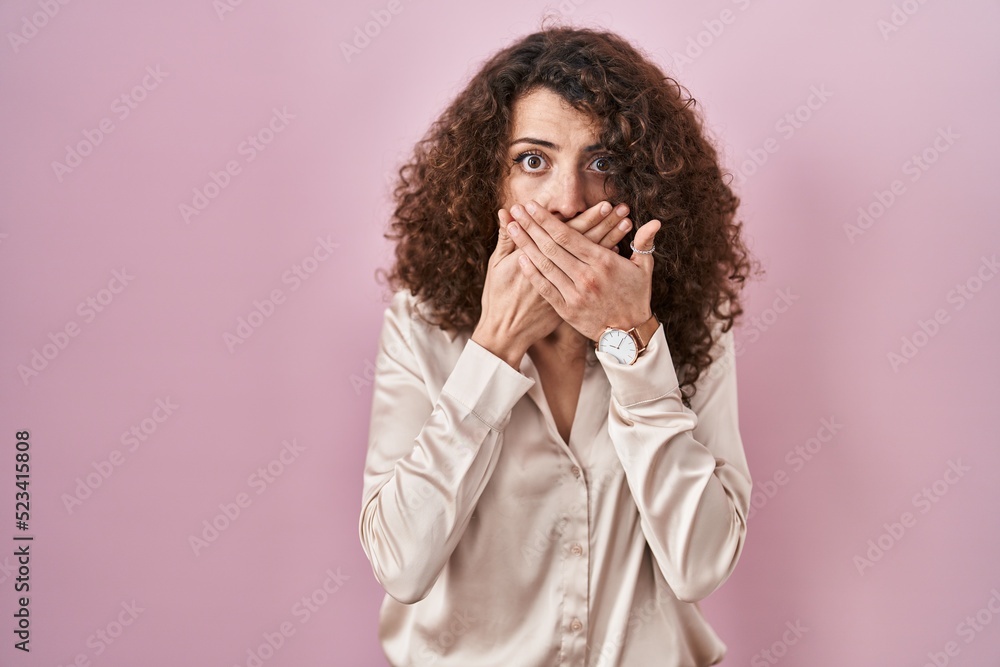 Hispanic woman with curly hair standing over pink background shocked covering mouth with hands for mistake. secret concept. Hispanic woman with curly hair standing over pink background shocked covering mouth with hands for mistake. secret concept.