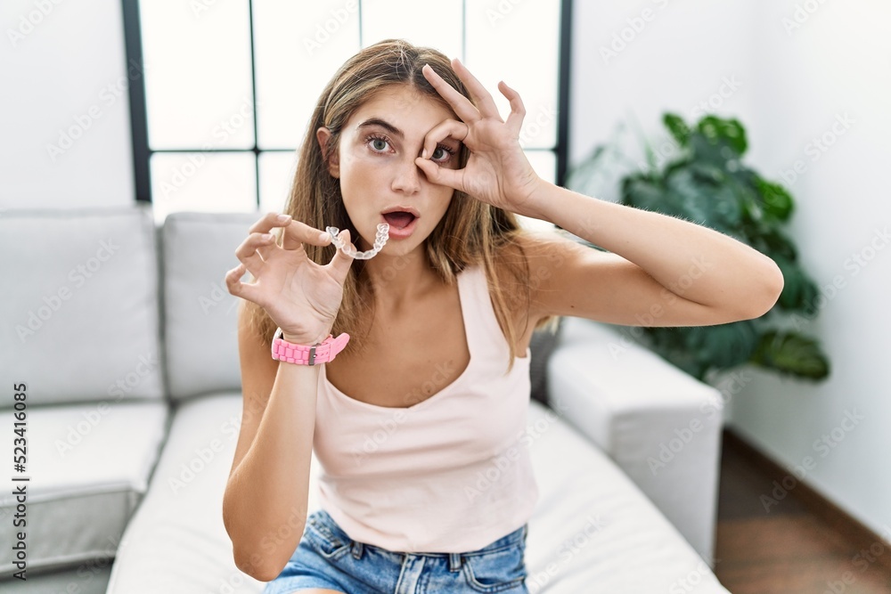 Young blonde woman holding invisible aligner orthodontic doing ok gesture shocked with surprised face, eye looking through fingers. unbelieving expression. Young blonde woman holding invisible aligner orthodontic doing ok gesture shocked with surprised face, eye looking through fingers. unbelieving expression.