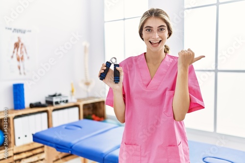 Young blonde woman working at pain recovery clinic holding hand strengthener pointing thumb up to the side smiling happy with open mouth