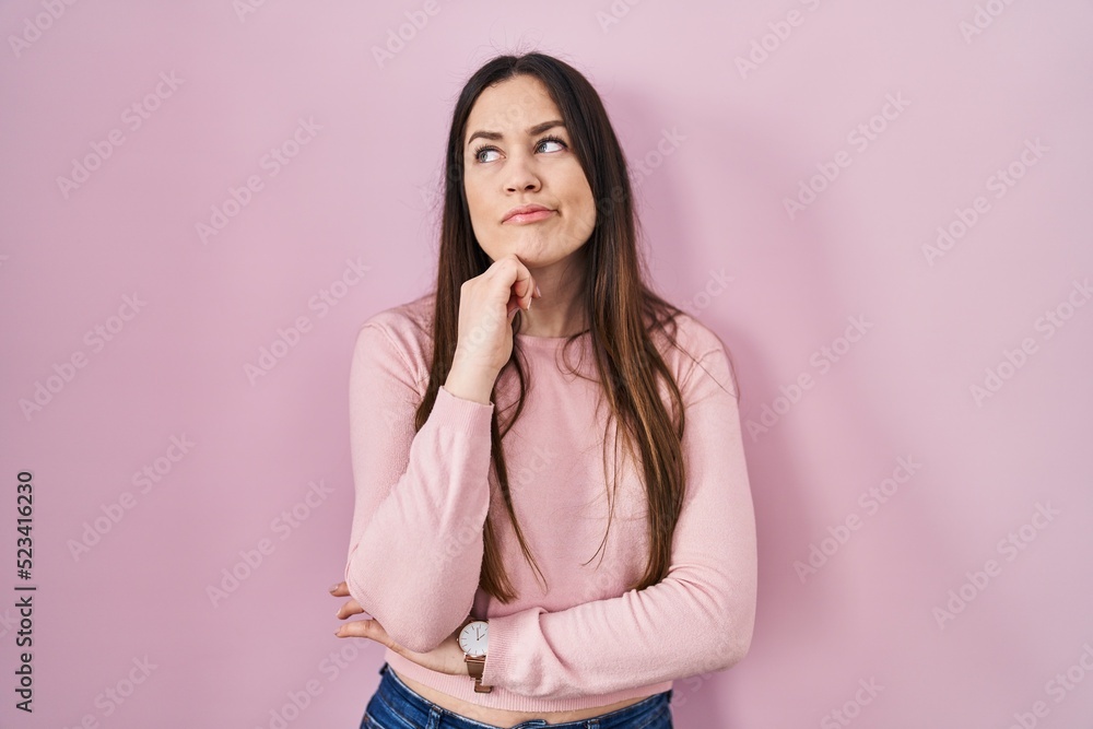 Young brunette woman standing over pink background with hand on chin thinking about question, pensive expression. smiling with thoughtful face. doubt concept.