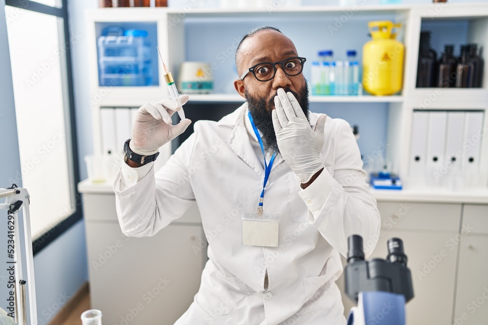 African american man working at scientist laboratory holding syringe covering mouth with hand, shocked and afraid for mistake. surprised expression African american man working at scientist laboratory holding syringe covering mouth with hand, shocked and afraid for mistake. surprised expression