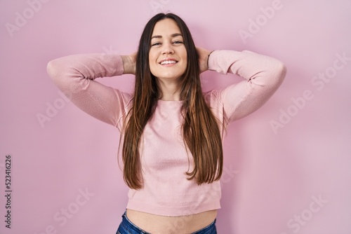 Young brunette woman standing over pink background relaxing and stretching, arms and hands behind head and neck smiling happy