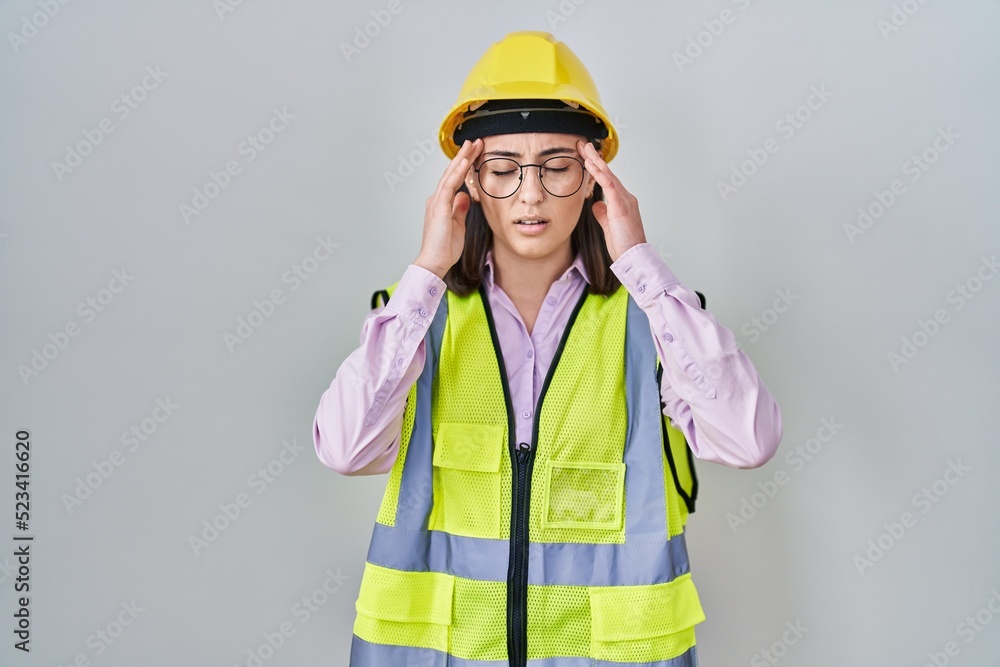 Hispanic girl wearing builder uniform and hardhat with hand on head for pain in head because stress. suffering migraine. Hispanic girl wearing builder uniform and hardhat with hand on head for pain in head because stress. suffering migraine.