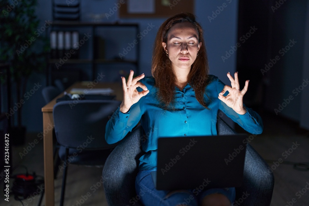 Brunette woman working at the office at night relax and smiling with eyes closed doing meditation gesture with fingers. yoga concept. Brunette woman working at the office at night relax and smiling with eyes closed doing meditation gesture with fingers. yoga concept.
