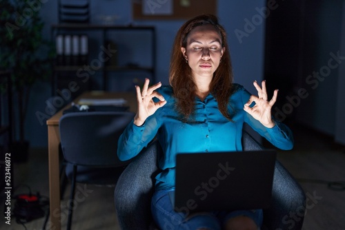 Brunette woman working at the office at night relax and smiling with eyes closed doing meditation gesture with fingers. yoga concept.