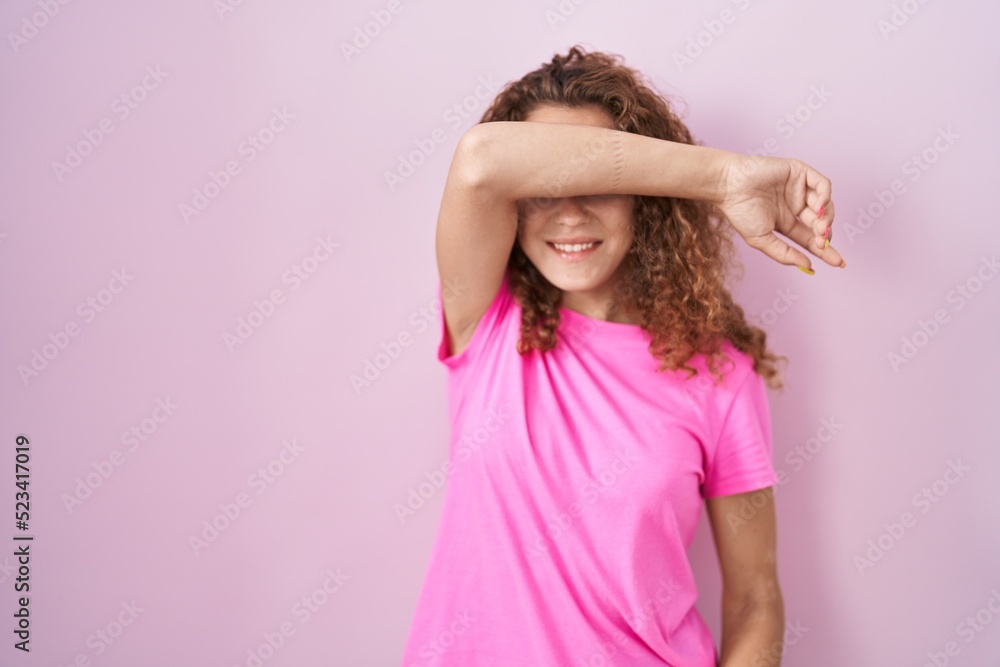 Young caucasian woman standing over pink background covering eyes with arm smiling cheerful and funny. blind concept. Young caucasian woman standing over pink background covering eyes with arm smiling cheerful and funny. blind concept.