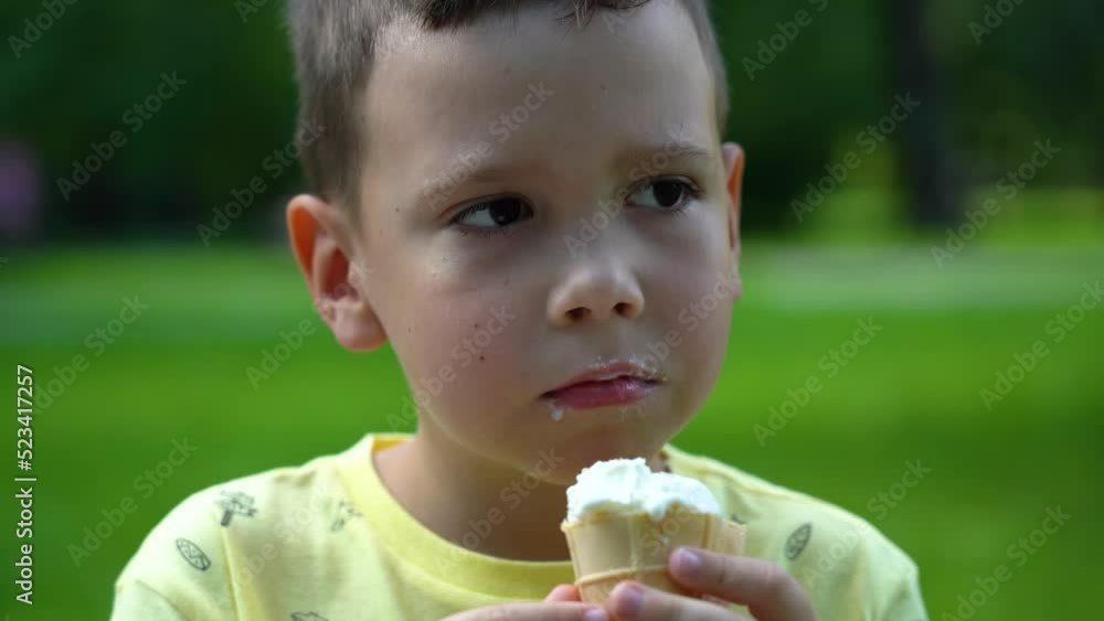 Caucasian boy eats ice cream on a sunny summer day under the trees in the park to satisfy his hunger and cool off. The boy eats delicious ice cream in the park among the green trees in the background Caucasian boy eats ice cream on a sunny summer day under the trees in the park to satisfy his hunger and cool off. The boy eats delicious ice cream in the park among the green trees in the background