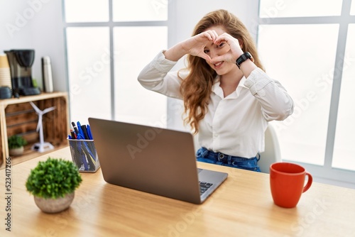 Young caucasian woman working at the office using computer laptop doing heart shape with hand and fingers smiling looking through sign