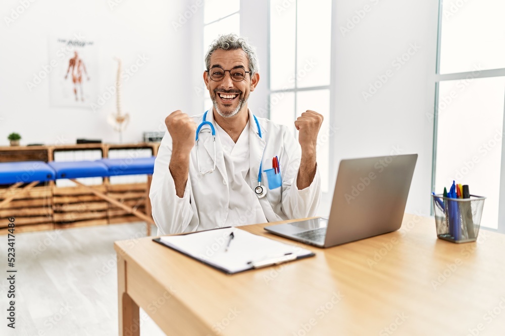 Middle age hispanic therapist man working at pain recovery clinic with laptop screaming proud, celebrating victory and success very excited with raised arms Middle age hispanic therapist man working at pain recovery clinic with laptop screaming proud, celebrating victory and success very excited with raised arms
