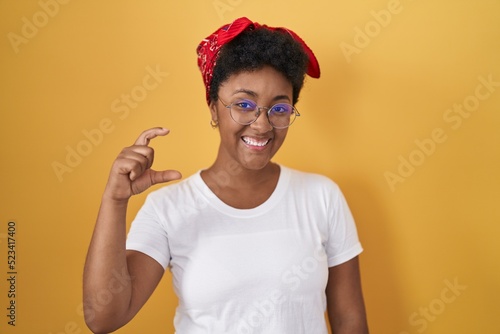 Young african american woman standing over yellow background smiling and confident gesturing with hand doing small size sign with fingers looking and the camera. measure concept.