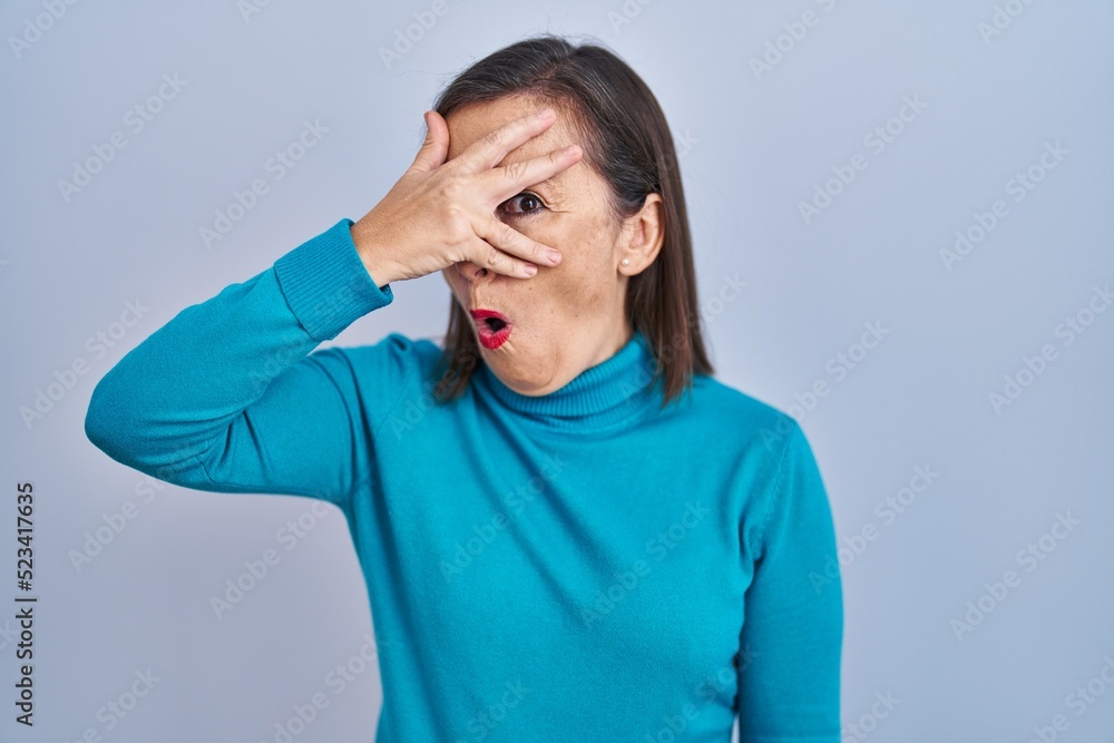 Middle age hispanic woman standing over isolated background peeking in shock covering face and eyes with hand, looking through fingers with embarrassed expression. Middle age hispanic woman standing over isolated background peeking in shock covering face and eyes with hand, looking through fingers with embarrassed expression.