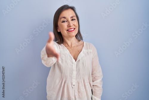 Middle age hispanic woman standing over blue background smiling friendly offering handshake as greeting and welcoming. successful business.