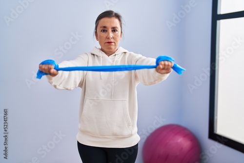 Middle age hispanic woman training arm resistance with elastic arm bands relaxed with serious expression on face. simple and natural looking at the camera.