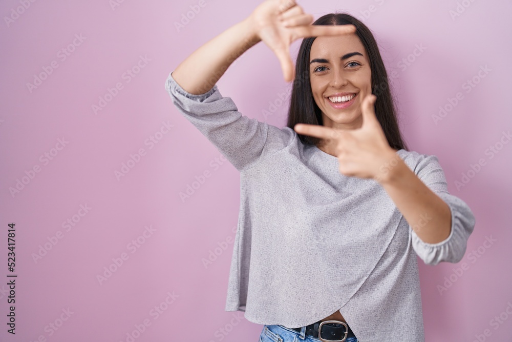 Young brunette woman standing over pink background smiling making frame with hands and fingers with happy face. creativity and photography concept. Young brunette woman standing over pink background smiling making frame with hands and fingers with happy face. creativity and photography concept.