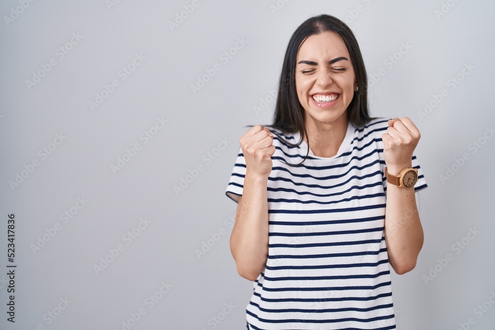 Young brunette woman wearing striped t shirt excited for success with arms raised and eyes closed celebrating victory smiling. winner concept. Young brunette woman wearing striped t shirt excited for success with arms raised and eyes closed celebrating victory smiling. winner concept.