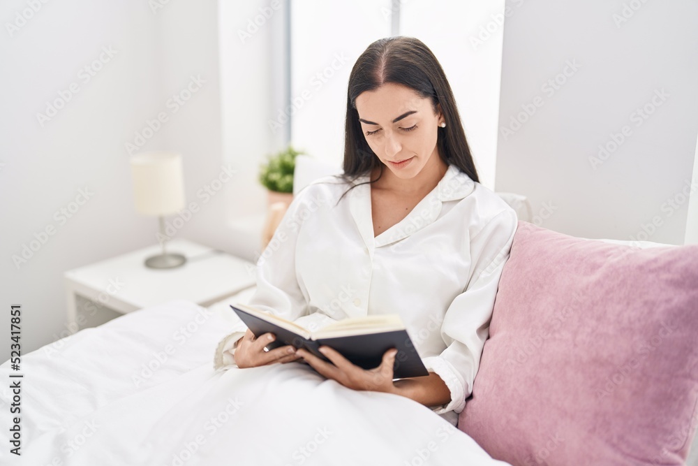 Young hispanic woman reading book sitting on bed at bedroom