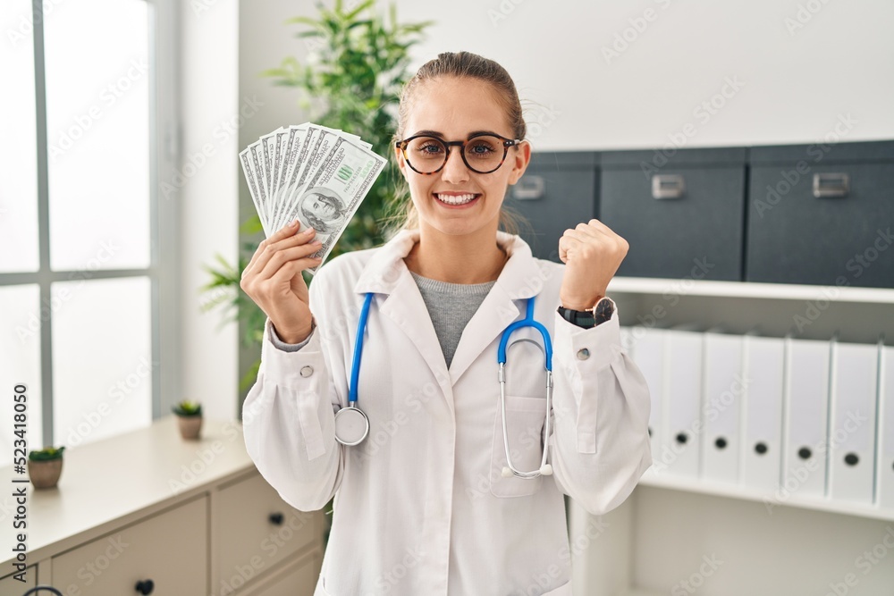 Young doctor woman holding dollars banknotes screaming proud, celebrating victory and success very excited with raised arm Young doctor woman holding dollars banknotes screaming proud, celebrating victory and success very excited with raised arm