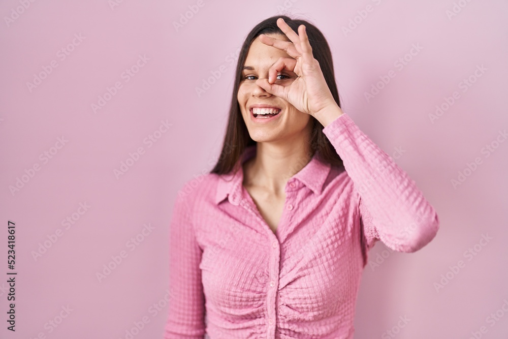 Young hispanic woman standing over pink background doing ok gesture with hand smiling, eye looking through fingers with happy face. Young hispanic woman standing over pink background doing ok gesture with hand smiling, eye looking through fingers with happy face.