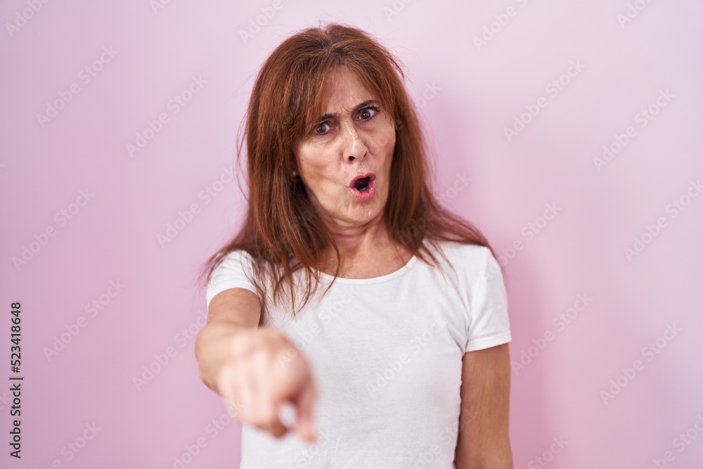 Middle age woman standing over pink background pointing displeased and frustrated to the camera, angry and furious with you Middle age woman standing over pink background pointing displeased and frustrated to the camera, angry and furious with you