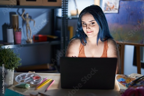 Young modern girl with blue hair sitting at art studio with laptop at night happy face smiling with crossed arms looking at the camera. positive person.