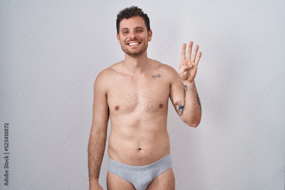 Young hispanic man standing shirtless wearing underware showing and pointing up with fingers number four while smiling confident and happy.