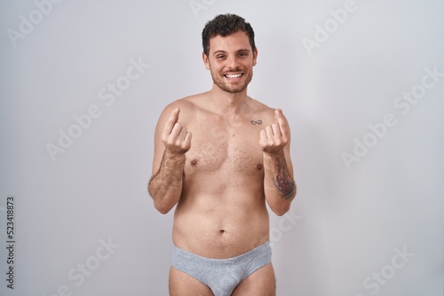 Young hispanic man standing shirtless wearing underware doing money gesture with hands, asking for salary payment, millionaire business