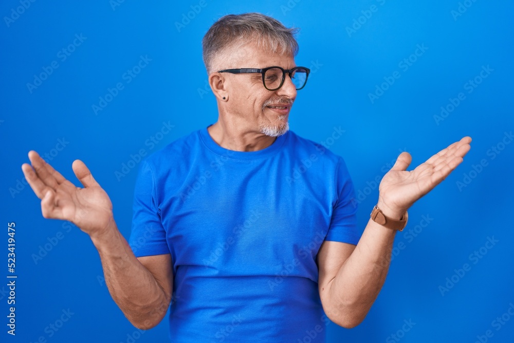 Naklejka premium Hispanic man with grey hair standing over blue background smiling showing both hands open palms, presenting and advertising comparison and balance